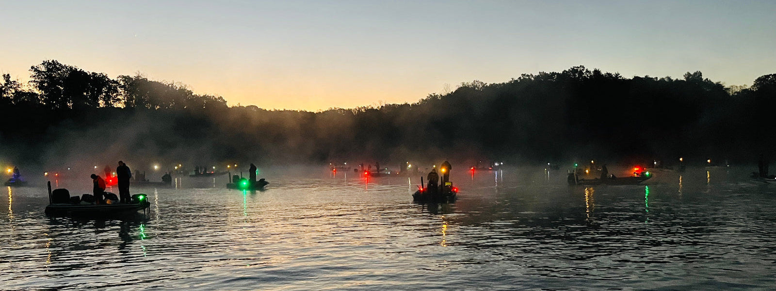 Bass fishing boats on a lake at dusk with lights on, surrounded by trees.