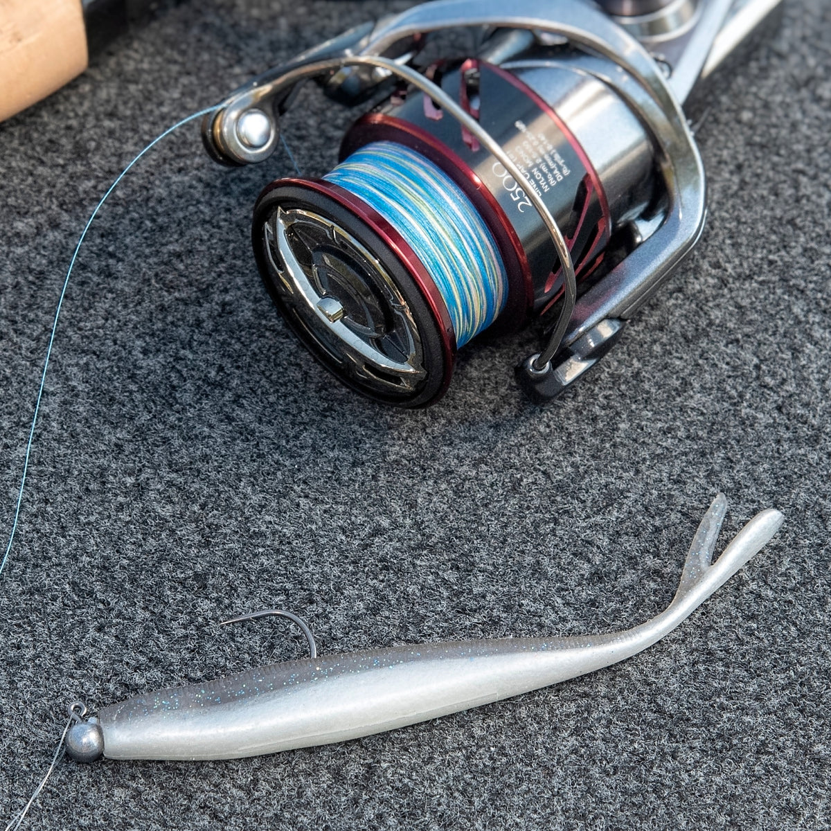 A close-up of a spinning fishing reel with blue and white line, placed on a dark textured surface next to a silver soft plastic swimbait lure.